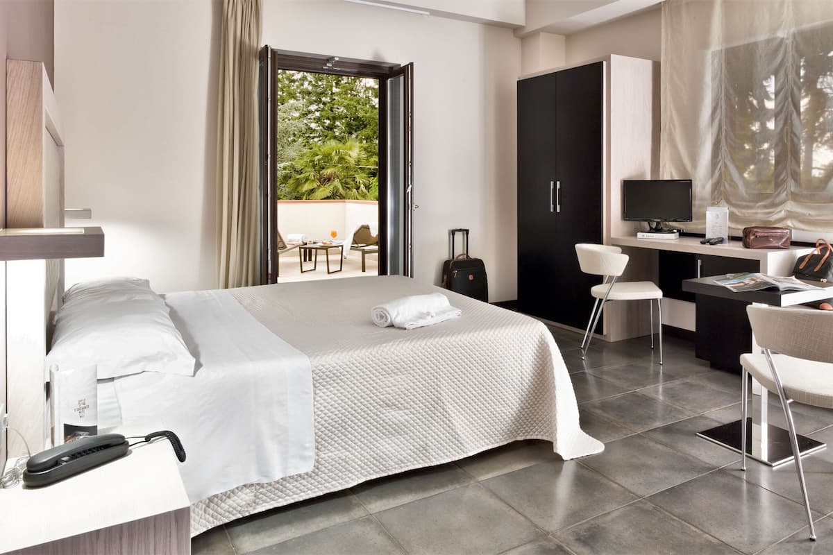 A hotel room with a bed, desk, chair, and a view of greenery outside.