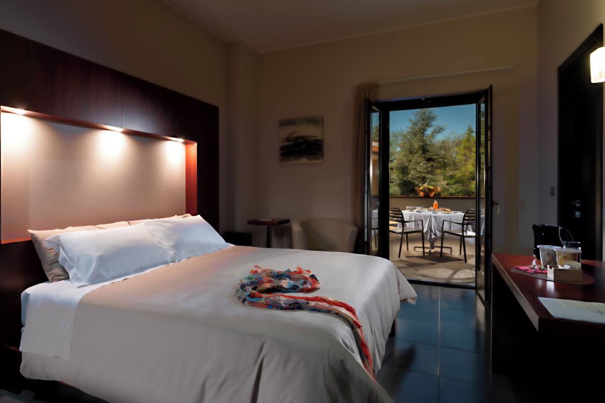 A hotel room with a bed, a small table with a chair, and a balcony with a table and chairs.