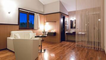 Una habitación de hotel moderna con suelo de madera, un sofá blanco, una mesita de noche con lámpara, una ventana con cortinas y un baño con espejo y lavabo.