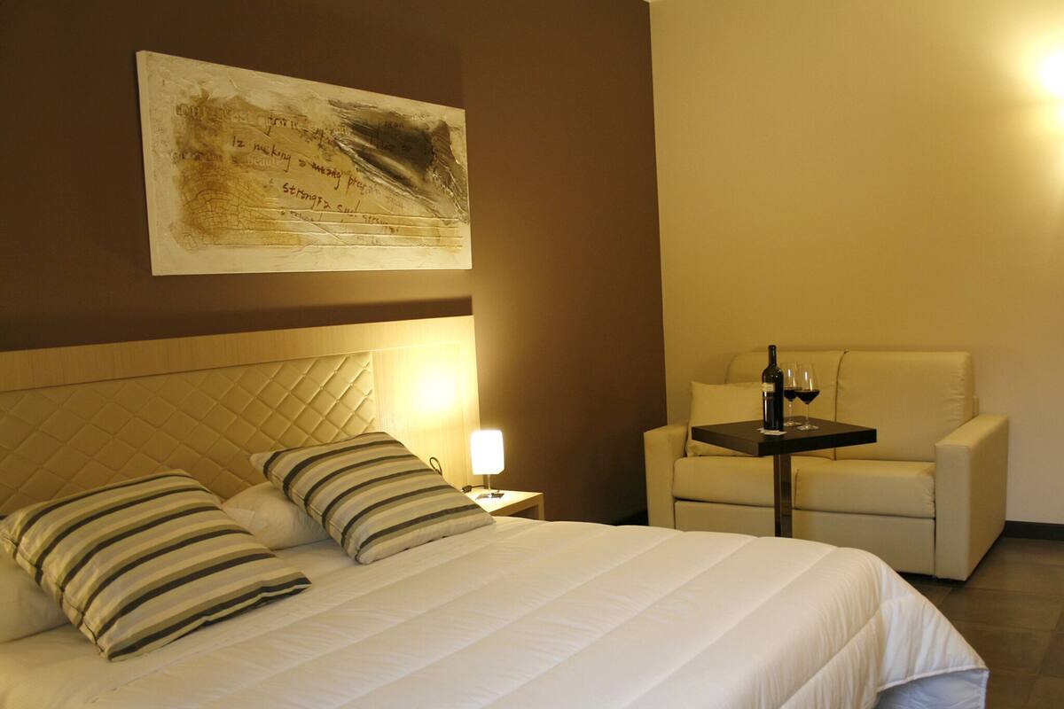 A hotel room with a bed, a chair, a small table with a bottle and glass, and a wall-mounted painting.