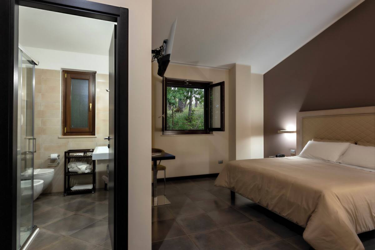 A bedroom with a bed, a window with a view of greenery, a bathroom with a toilet and sink, and a small table with chairs.