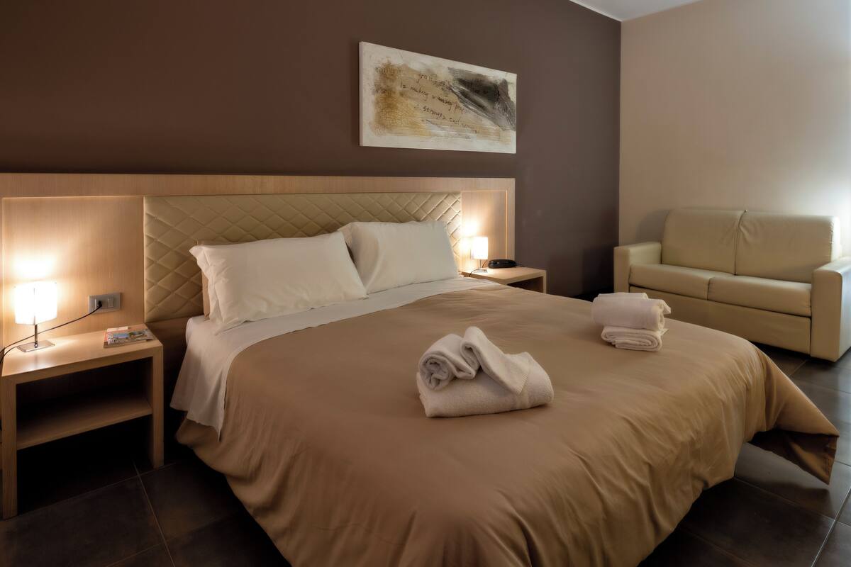 A hotel room with a large bed, two bedside tables, a nightstand, a sofa, and a painting on the wall.