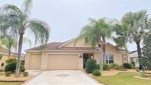 A single-story house with a beige exterior, a garage, and a front yard with palm trees and shrubs. - Spectacular Hot Tub w/ Pool Home on Beautiful Golf Course View,BBQ, & WiFi! (The Villages)