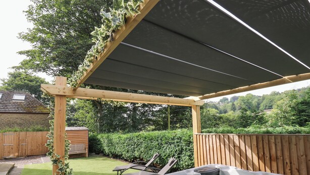 A hot tub under a wooden pergola with a view of a garden and a wooden fence.