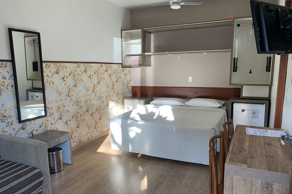 Ein Hotelzimmer mit Bett, Schreibtisch, Stuhl, einer kleinen Mülltonne, einem Spiegel und einem Fernseher.