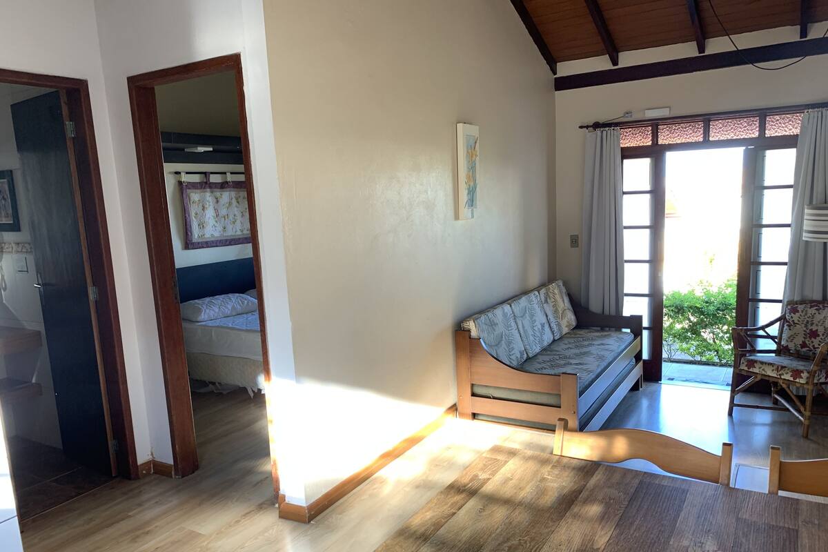 Ein Zimmer mit einer hölzernen Sitzbank, einem Sofa mit gemusterten Kissen, einem Bett mit floralem Überwurf und Blick auf viel Grün durch große Fenster.