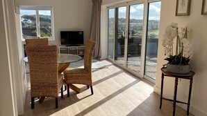 A spacious living room with a large sliding glass door leading to a balcony, wicker chairs, a small table with a potted orchid, and a flat-screen TV on a stand. - Spacious Retreat Private Hot Tub Sea/Downs views/patio 10min walk to Beach/Train (seaford)