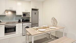 A modern kitchen with white cabinets, a built-in oven, microwave, and refrigerator. There is a dining table set with plates, glasses, and cutlery. - Sunset On Hill 3 (Cape Town)
