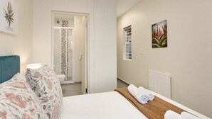 A hotel room with a bed, a bathroom with a shower, and a hallway with a radiator and artwork. - Sunset On Hill 3 (Cape Town)
