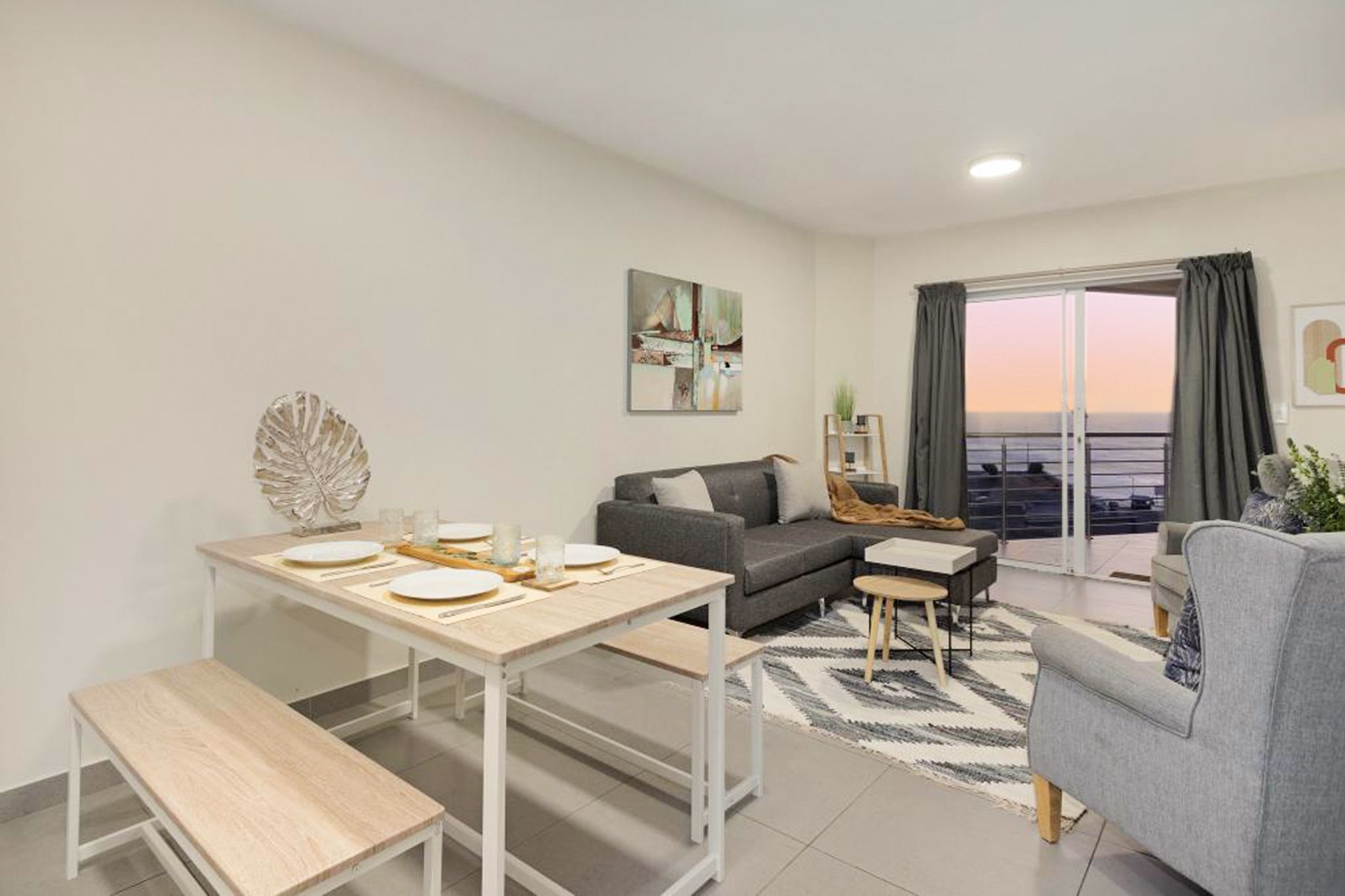 A modern living room with a dining area, a gray sofa, a coffee table, and a view of the sea through large windows.