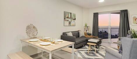 A modern living room with a dining area, a gray sofa, a coffee table, and a view of the sea through large windows.