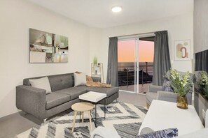 A modern living room with a grey sofa, a small coffee table, and a view of the sunset through the balcony. - Sunset On Hill 3 (Cape Town)