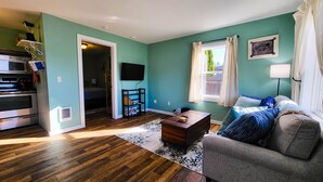 A living room with a wooden floor, a grey sofa, a coffee table, and a TV mounted on the wall. - Bay Street Retreat - Waldport (Waldport)