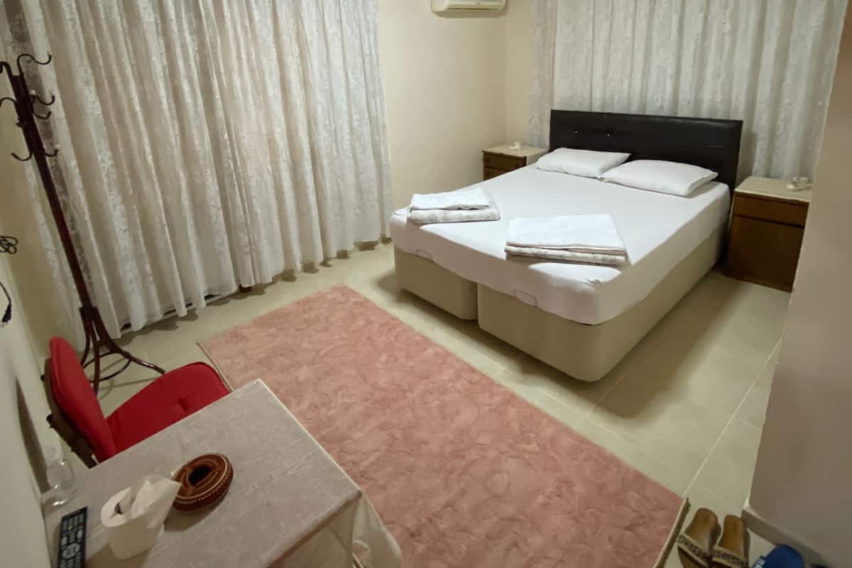 Een slaapkamer met een bed, een tafel, een stoel, een rode stoel, een roze vloerkleed, een airconditioner en een plafondlamp.