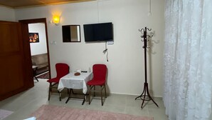 Habitación con ventilador de techo, televisor de pantalla plana montado en la pared, una mesa de comedor con dos sillas, perchero y un ventanal con cortinas.