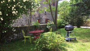 Un bâtiment en pierre avec une porte rouge, une table et des chaises rouges, un barbecue et un petit arbuste.