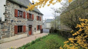 Un bâtiment en pierre avec des volets rouges et un chemin pavé menant à une zone herbeuse parsemée de fleurs jaunes.