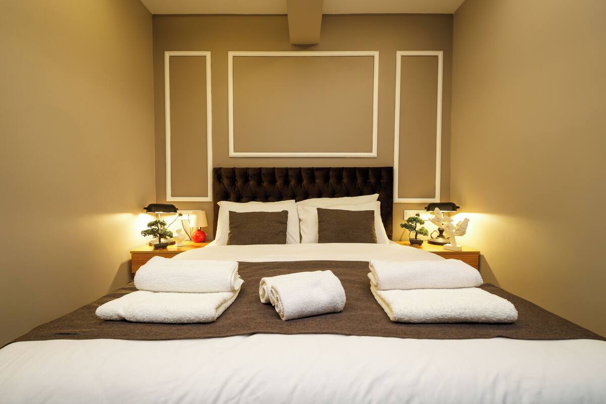 A neatly made bed with white linens and a brown throw blanket, flanked by two bedside tables with lamps and decorative items.