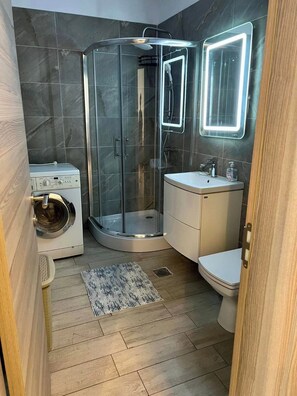 A modern bathroom with a glass shower enclosure, a washing machine, a sink, and a toilet. - Deluxe Alezzi (Navodari)