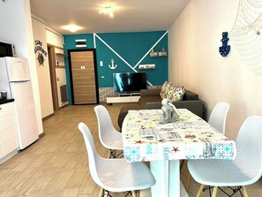 A modern living room with a teal accent wall, a flat-screen TV, a dining table with white chairs, and a kitchen area with a white refrigerator.