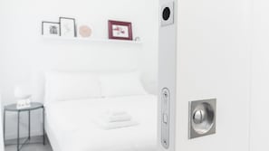 Una habitación de hotel moderna con una cama blanca, una mesita de noche pequeña y un estante con marcos que contienen fotografías.
