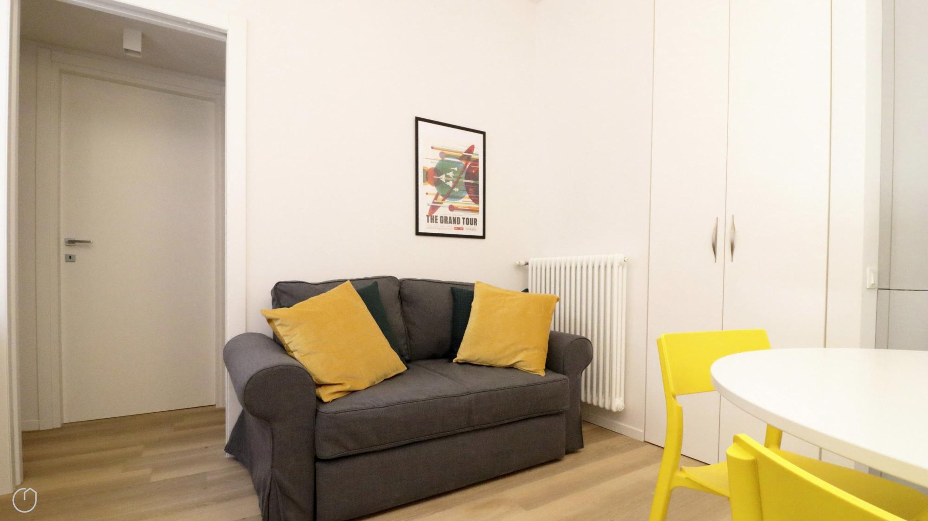 Una habitación con un sofá gris, dos cojines color mostaza, un radiador blanco, una mesa blanca y una silla amarilla.