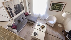 Uma sala de estar moderna com um sofá modular cinza, uma poltrona branca e uma pequena mesa branca. Há livros e um abajur sobre a mesa.