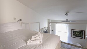 Quarto de hotel com uma cama grande, um ventilador de teto, uma janela com cortinas e uma moldura com um quadro na parede.