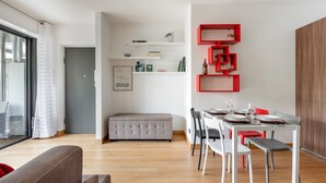 Ein moderner Essbereich mit einem Holztisch, gedeckt für vier Personen, umgeben von weißen Stühlen. Eine graue Bank steht an der Wand, und darüber an der Wand ist ein rotes Regal angebracht.