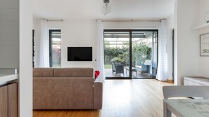 Una sala de estar moderna con un sofá, una mesa de comedor y un televisor montado en la pared. Hay una puerta corrediza de vidrio que da a un patio exterior con una mesa y sillas.
