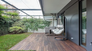 A modern outdoor patio with a wooden deck, a hanging chair, and a glass door leading to a garden area with greenery.