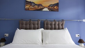 Une chambre à coucher avec un lit, deux lampes de chevet, une tête de lit ornée de coussins capitonnés, un tableau représentant un paysage urbain et une prise de courant sur le mur.