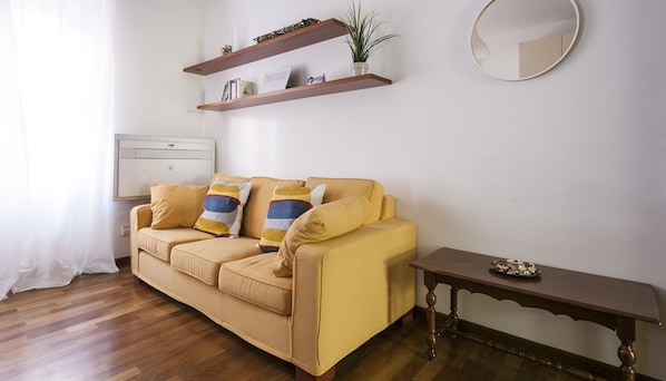 A living room with a yellow sofa, two cushions, a small table, a mirror, and two wooden shelves with decorative items. - Cirillo 14 - Apartment Milan (Milan)