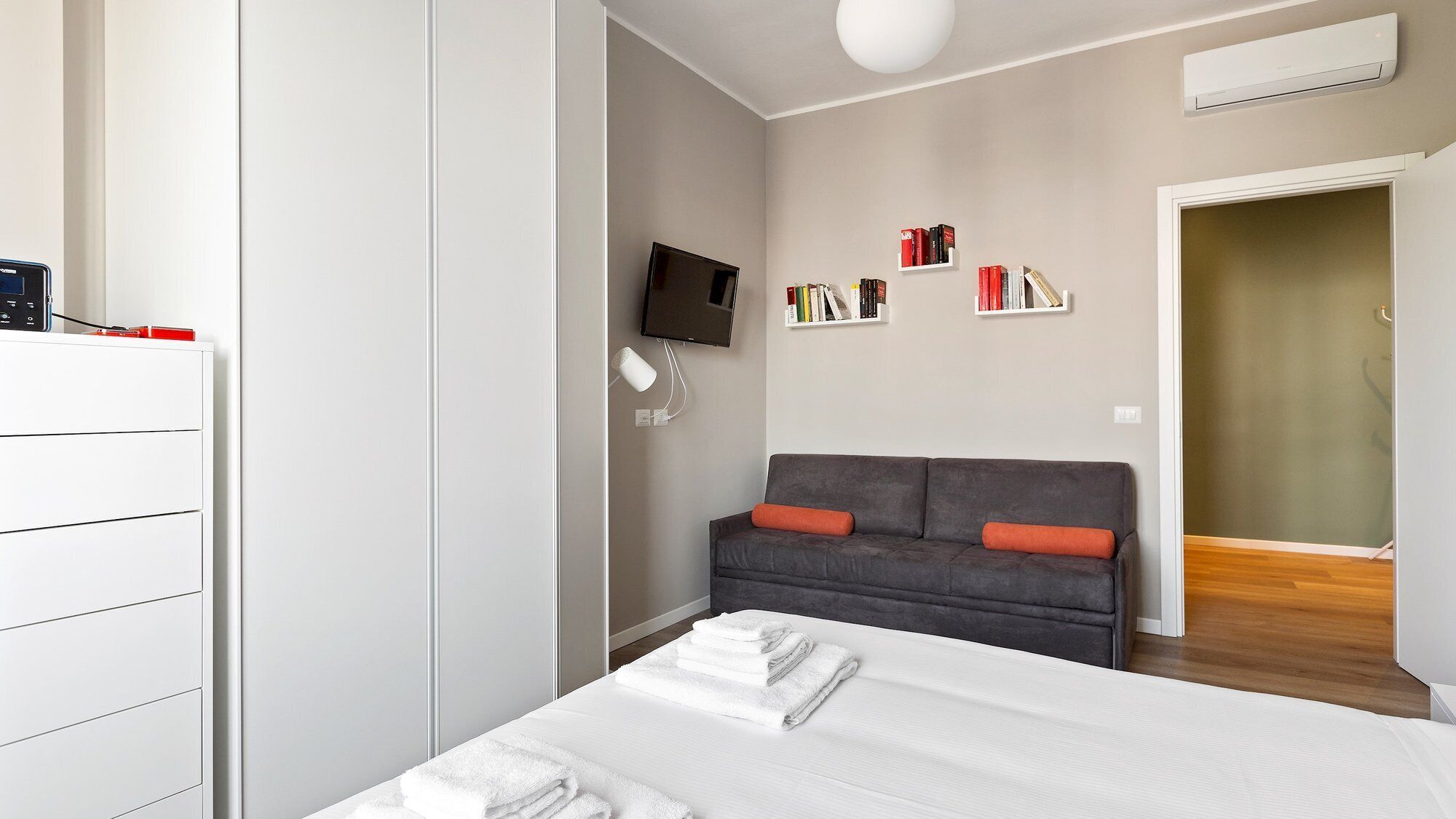 Een moderne slaapkamer met een bed, een grijze bank met oranje kussens, een televisie en een boekenplank vol boeken.