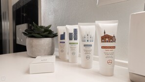 A row of skincare products on a white surface, including body wash, shampoo, and body cream, with a potted plant and a box in the background.