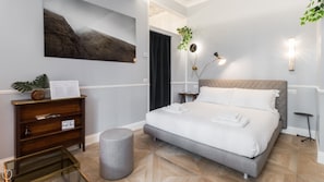 A modern hotel room with a large bed, a wooden dresser, a round ottoman, and a wall-mounted clock.
