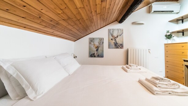 A bedroom with a wooden sloped ceiling, a bed with white linens, two wall-mounted paintings, a radiator, and a wooden dresser.