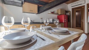 A dining area with a wooden table set for four, white plates, bowls, and wine glasses. There are white chairs, a red refrigerator, and wooden cabinets in the background.