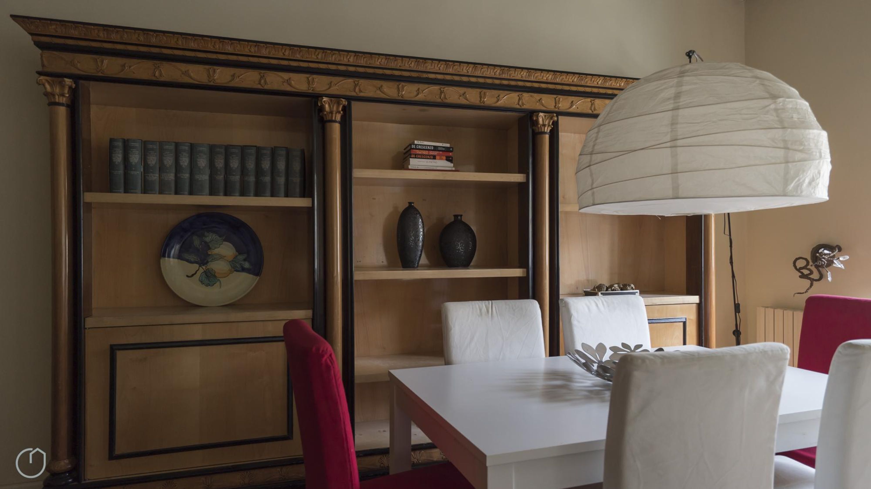 A dining area with a white table and chairs, a decorative vase, and a bookshelf with books and a plate.