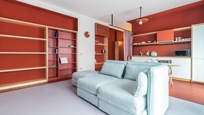 Een moderne woonkamer met een lichtblauwe bank, rode kasten en een keukenhoek met een magnetron en waterkoker.