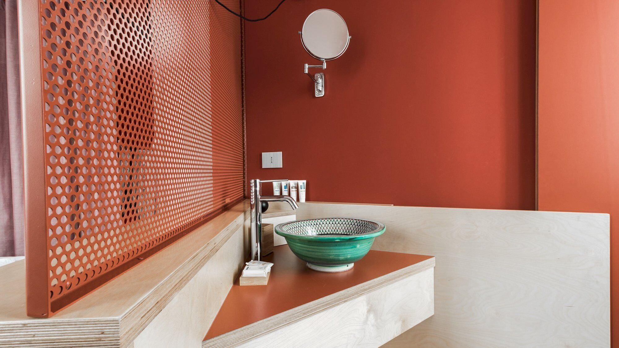 A bathroom with a wooden vanity, a green glass bowl sink, a mirror, and a perforated metal partition.