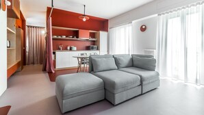 Een moderne woonkamer met een grijze hoekbank, een kleine eettafel en een keuken met een magnetron en waterkoker.