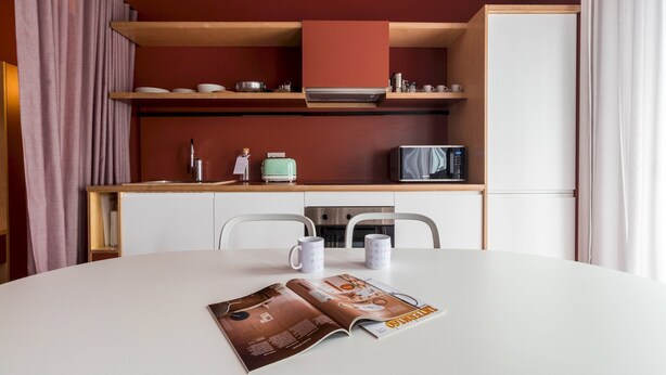 Een moderne keuken met witte kastjes, een houten aanrecht, een magnetron en een waterkoker. Er staat een eettafel met twee stoelen en een tijdschrift erop.