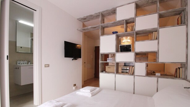 A bedroom with a bed, a TV mounted on the wall, and a built-in shelving unit with books and decorative items. - Bligny 39 B (Milan)