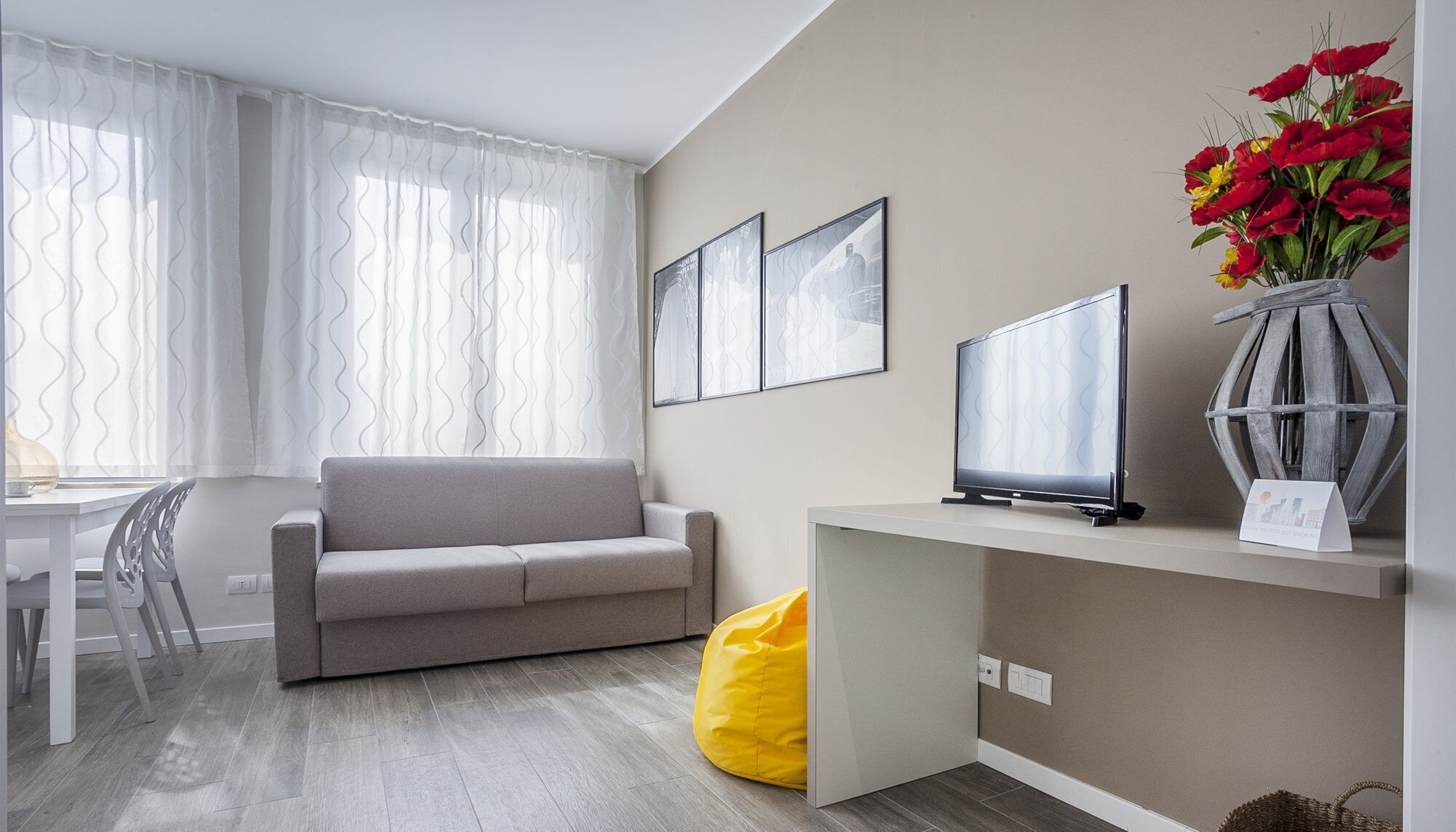 Ein modernes Wohnzimmer mit einem grauen Sofa, einem Fernseher auf einem Schrank, einem gelben Sitzsack und einer Vase mit roten Blumen.