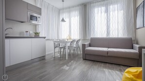 A modern kitchen with a built-in microwave, a dining area with a table and chairs, and a grey sofa in a living area with large windows and curtains. - Italianway Easy - Cadorna 10 Mono A (Milan)