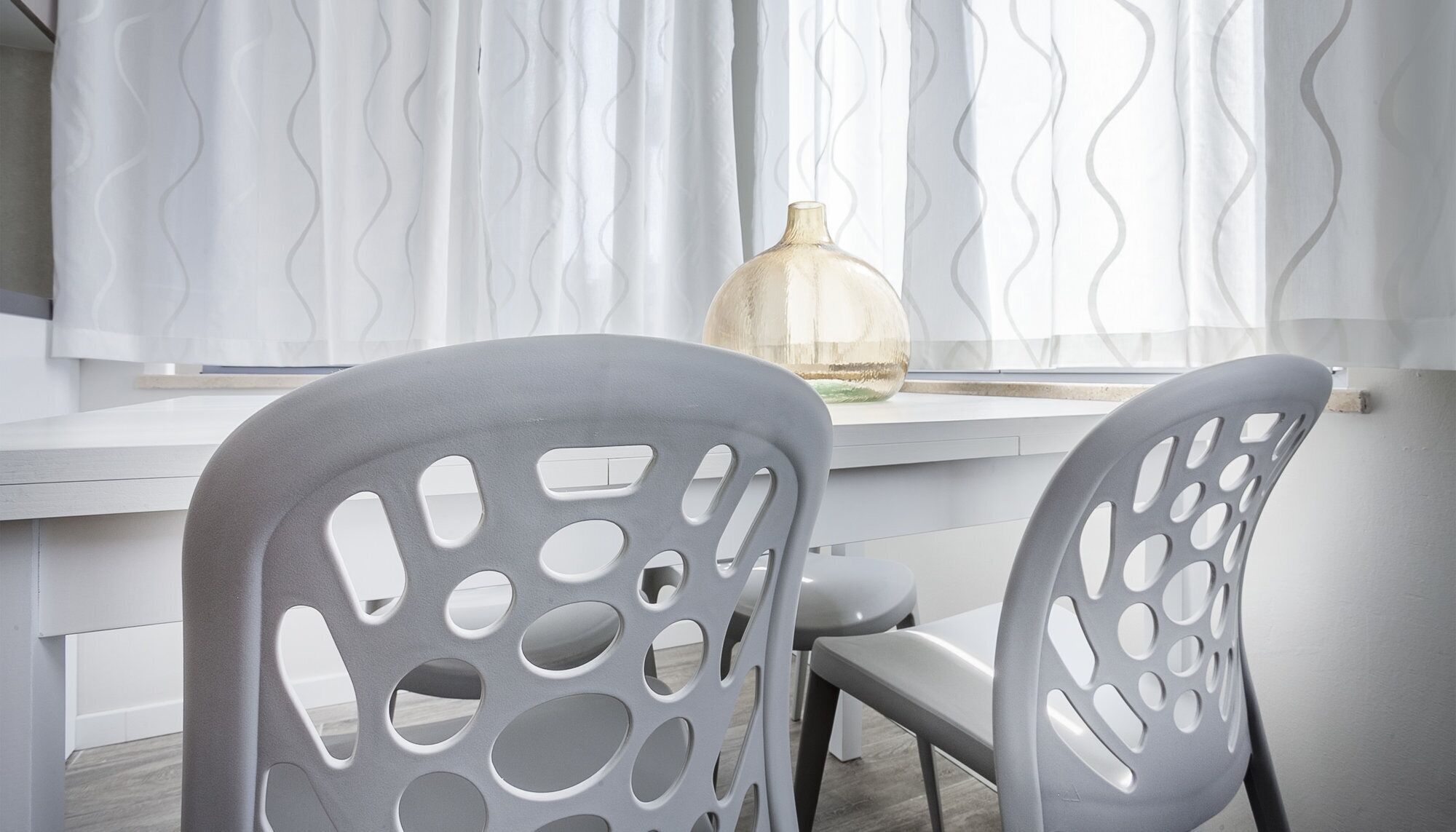 A modern room with a white desk, two grey chairs with circular cutouts, a clear vase on the desk, and wavy white curtains.