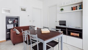 A modern living room with a wooden dining table, grey chairs, a brown sofa, a bookshelf, and a television. - Italianway Easy - Cerano 15 (Milan)