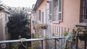 Une rue étroite en milieu urbain, bordée d’immeubles très rapprochés, avec des balcons dotés de garde-corps en métal et un arbre au premier plan.