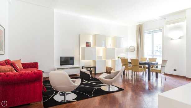 A modern living room with a red sofa, white armchairs, a dining area with yellow chairs, and a TV on a white stand. - Italianway Easy - Cristoforo Colombo 1 (Milan)